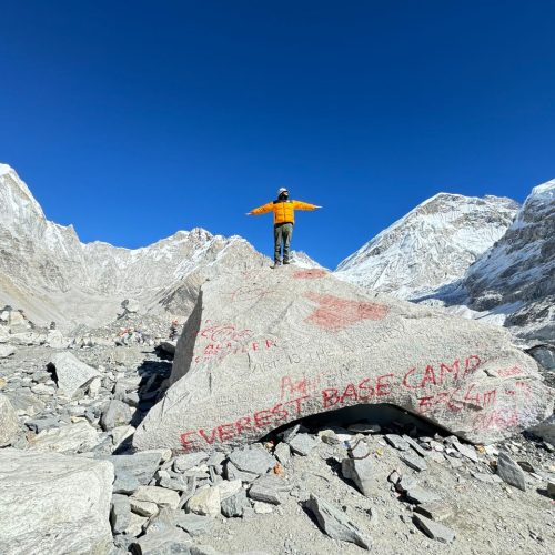 A mountaineer stands on a rock at Everest Base Camp, surrounded by snowy peaks.